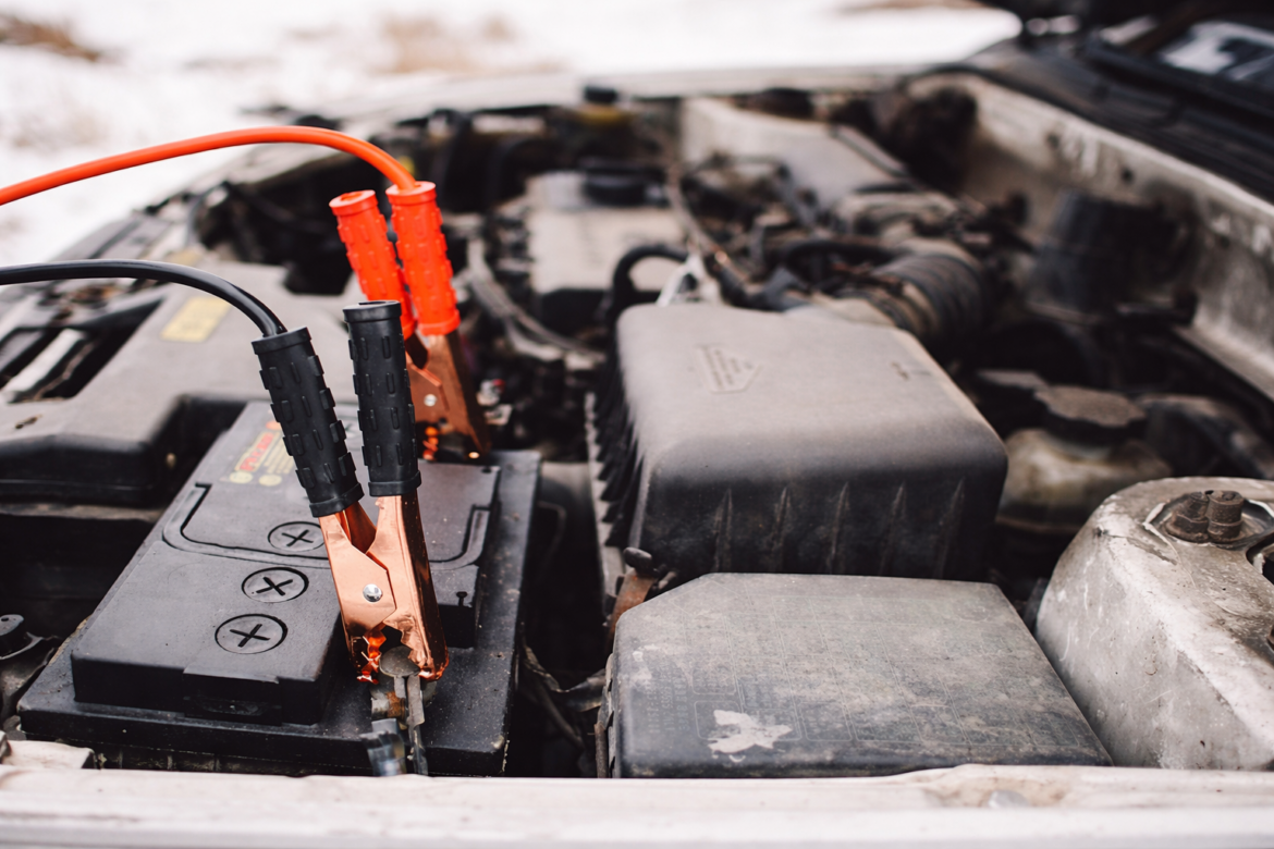 Nahaufnahme einer Autobatterie im Motorraum mit angeschlossenen Starthilfekabeln bei winterlichen Bedingungen.