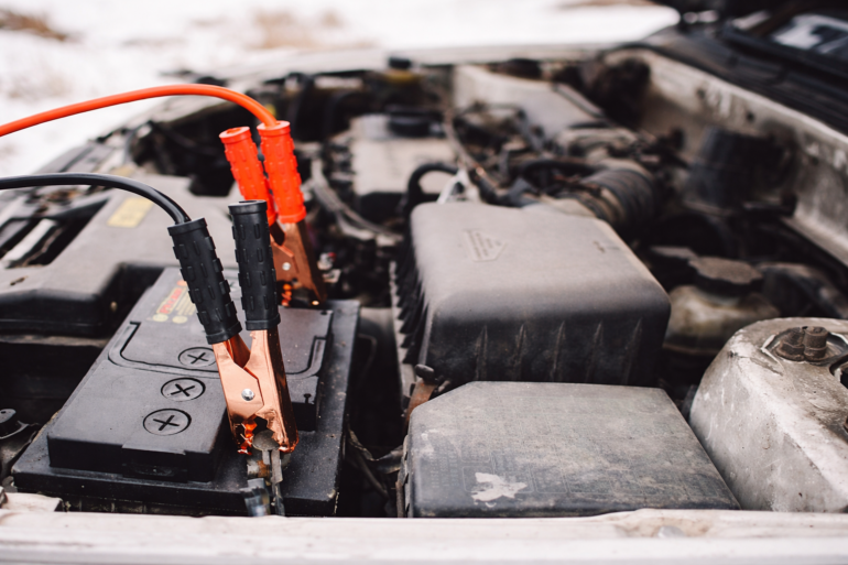 Nahaufnahme einer Autobatterie im Motorraum mit angeschlossenen Starthilfekabeln bei winterlichen Bedingungen.